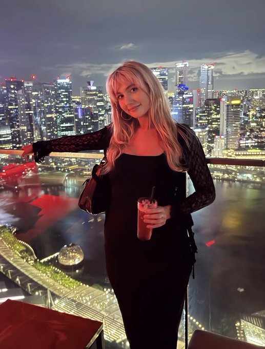 A blonde woman in a black lace dress poses on a rooftop at night, holding a cocktail with a stunning city skyline of illuminated skyscrapers and a waterfront visible behind her, fitting the post's announcement about attending @token2049 with collaboration opportunities.