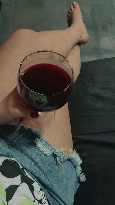 A first-person view of a person in denim shorts sitting on a dark surface, holding a glass of red wine with bare feet visible.