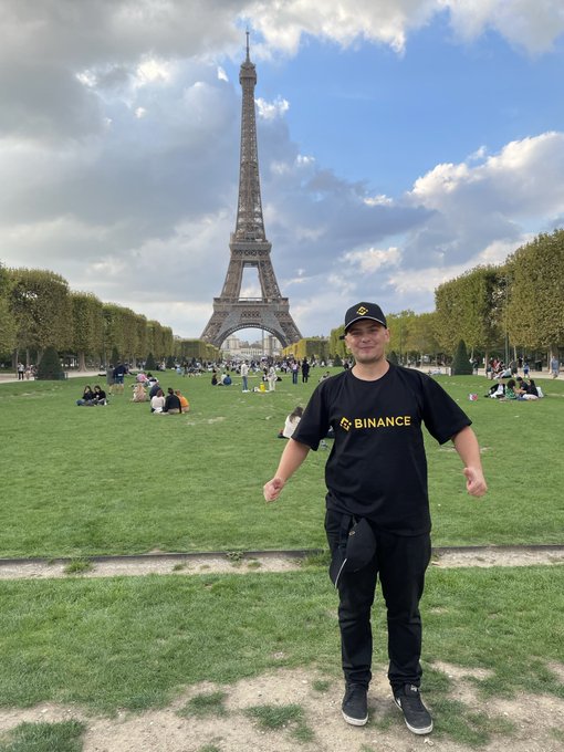 A man wearing a black cap and a black t-shirt branded with the yellow 'BINANCE' logo stands in front of the Eiffel Tower in Paris.