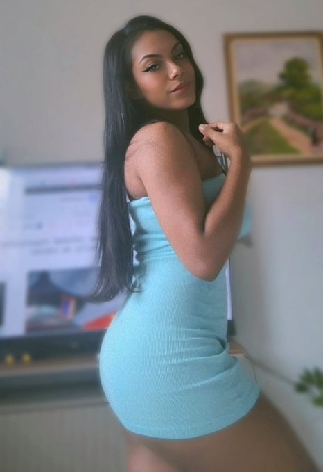 A woman with long black hair posing in a light blue strapless dress, looking over her shoulder in an indoor setting with a framed painting on the wall.