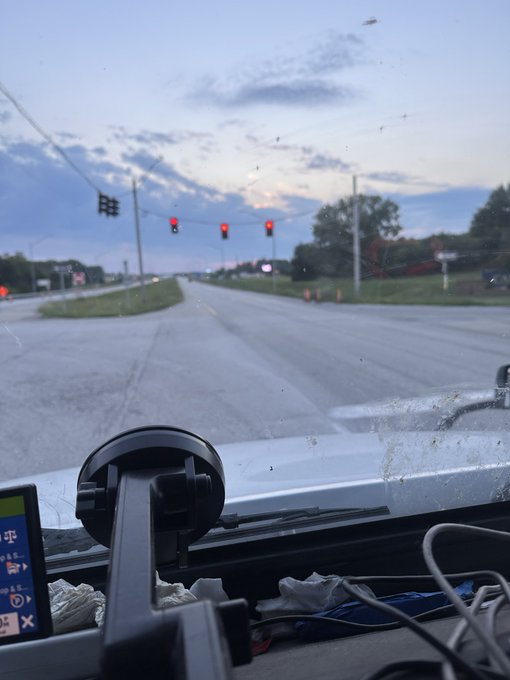 A view from inside a vehicle shows red traffic lights at an intersection during dusk, with a phone mount on the dashboard and bugs visible on the windshield.