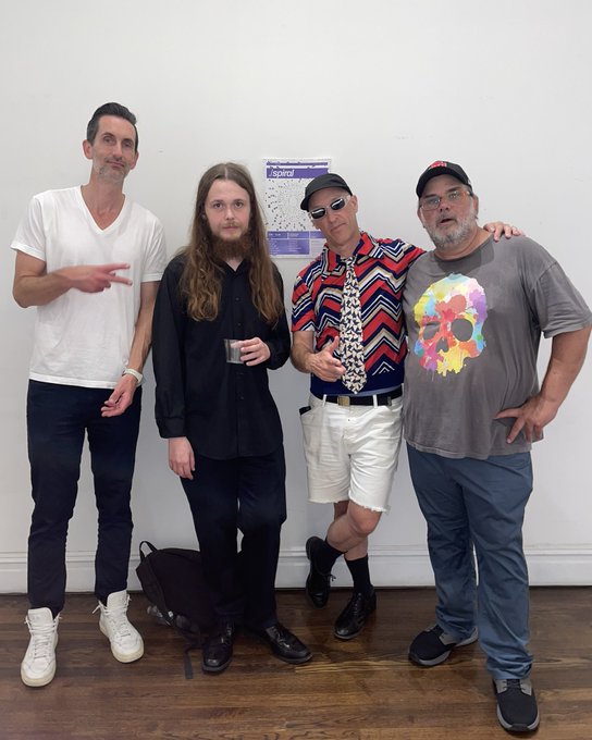 Four men pose together for a photo in front of a white wall with a small calendar poster; they are dressed casually with one wearing a distinctive red patterned shirt and tie.