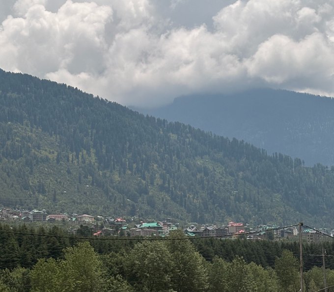 A scenic view of a small town nestled at the base of densely forested mountains under a cloudy sky, with power lines visible in the foreground.