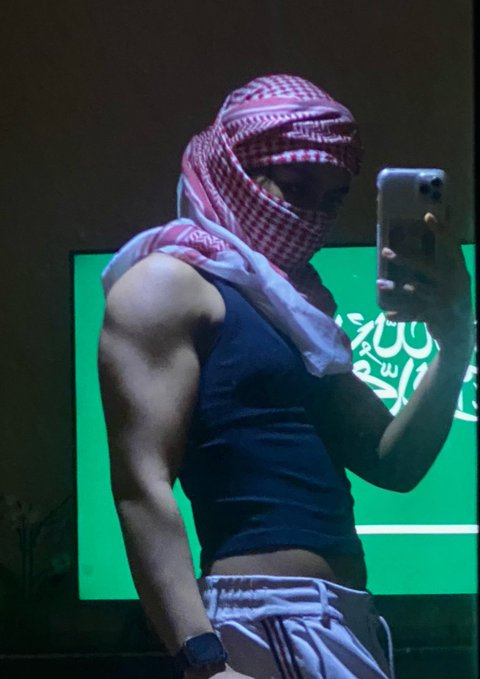 A muscular person wearing a red and white keffiyeh covering their face and head, taking a mirror selfie with a smartphone while wearing a black tank top and white pants. Behind them, a green flag with the Saudi Arabian flag design and Arabic text is visible. The post hashtag "#طخ" (meaning "spray" or "splash" in Arabic slang) relates to the image through the spray-painted aesthetic of the pose and the flag prominently displayed in the background.