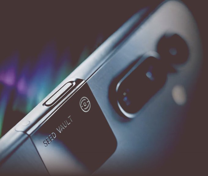 A close-up product photograph of a smartphone featuring a dual-camera module and a 'SEED VAULT' label with a small logo on its metallic body, set against a blurred iridescent background.