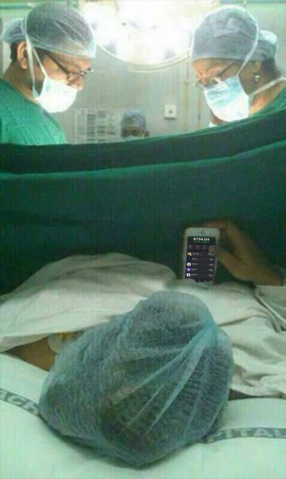 A person lying in a hospital bed under a surgical cap and drape shows a smartphone displaying a crypto portfolio worth $734.04, while two surgeons in masks look on in the background.
