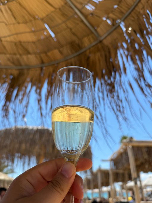 A hand raises a champagne flute of sparkling wine in a toast beneath a thatched beach umbrella, with tropical cabanas and lounge chairs visible in the sunny background. The celebratory vacation scene pairs perfectly with the post's announcement of an upcoming "data glow up," toasting to success while soaking up the SPF from the beach.