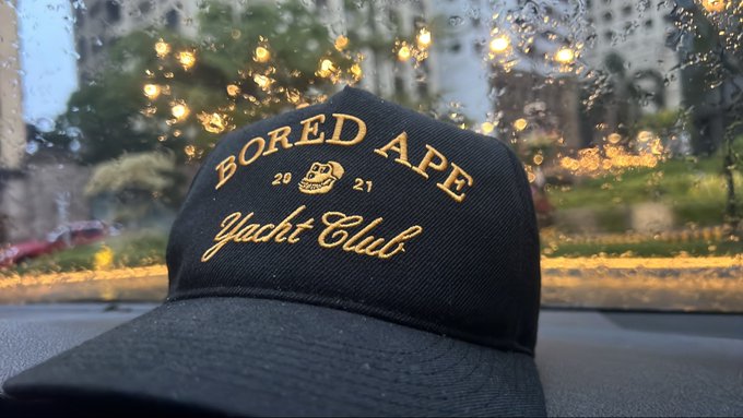 A black baseball cap rests on a car dashboard with rain-speckled windows in the background, featuring gold embroidered text reading "BORED APE Yacht Club" with "20 21" flanking a small ape logo. The cap serves as branded merchandise for the Bored Ape Yacht Club NFT collection, prominently displaying the community's name in a classic varsity-style font.