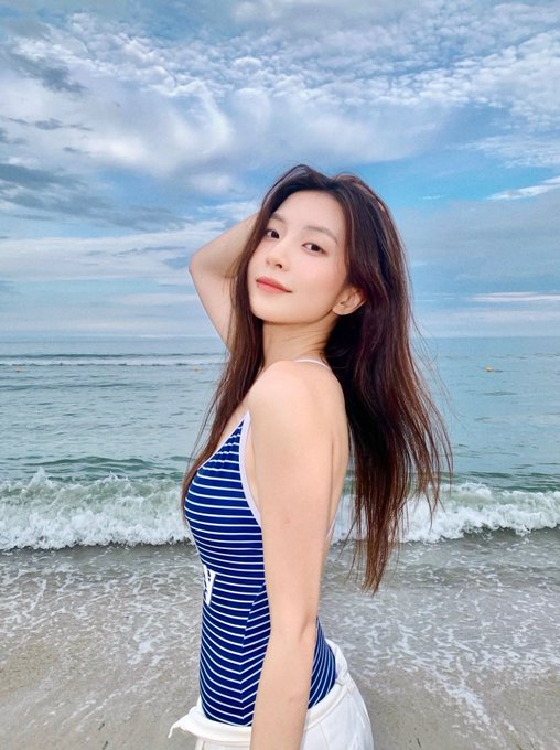 A young woman with long brown hair poses at the beach wearing a blue and white striped swimsuit top and white pants, with the ocean and a cloudy sky behind her.