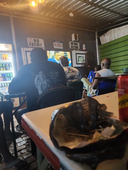 A group of patrons watch a soccer match on TV at a local eatery featuring "NO CREDIT," "NO IDLE SITTING," and "NO CHAINS" warning signs, with a black bag and red soda can on the foreground table, matching the caption's scene of supporting France during a game.