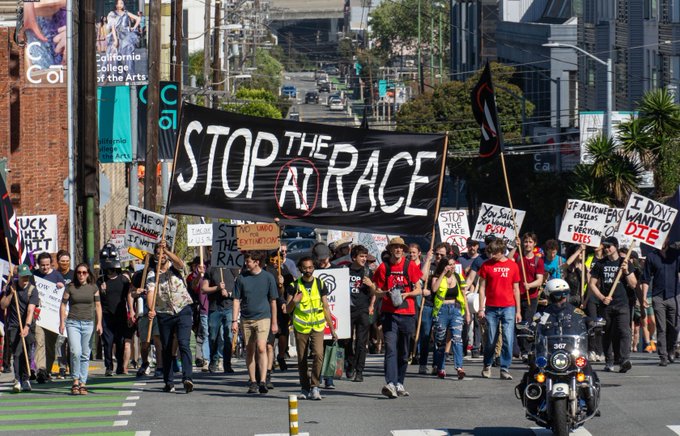 A street protest with marchers carrying a large black banner reading 'STOP THE AI RACE', along with other signs opposing AI development.