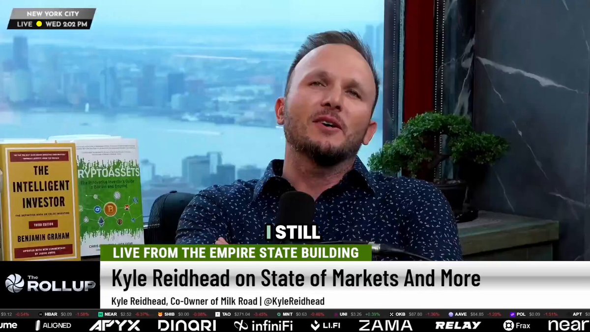 The image shows a man sitting at a desk with a book titled "The Intelligent Investor" by Benjamin Graham, which he is reading while wearing a blue shirt. The background of the image features a city skyline, suggesting that they are located in New York City.