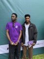 A small, blurry photo of two men standing side by side outdoors, one wearing a purple shirt and the other in a dark jacket.