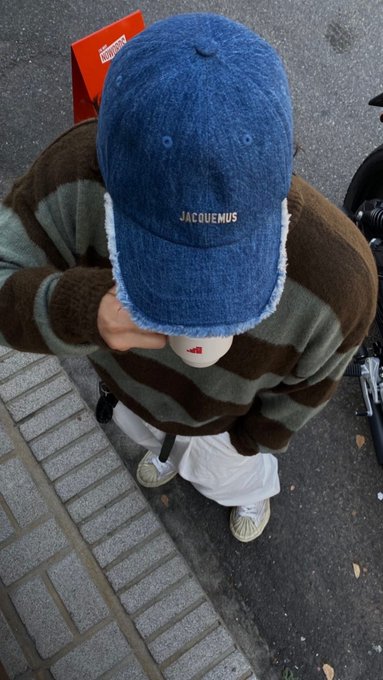 A person wearing a blue 'JACQUEMUS' cap and a brown and green striped sweater stands on a brick sidewalk looking down, with the top of their head and cap filling the frame.