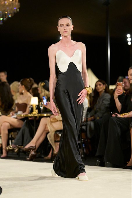 A fashion model walks down a runway wearing a strapless black satin gown with a white sweetheart neckline bodice, accessorized with a gold bracelet.