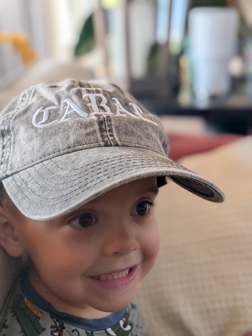 A young boy smiles brightly while wearing a faded baseball cap embroidered with the word "CABAL," perfectly playing into the pun of his dad joking that he "joined the Cabal" on a Saturday morning.
