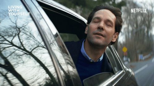The image features a man sitting inside a car, looking out of the window with his eyes closed. The scene appears to be set in an urban environment, as there are trees visible outside the car window. The caption for this image reads "living with yourself", which is likely related to the movie "Living With Yourself".