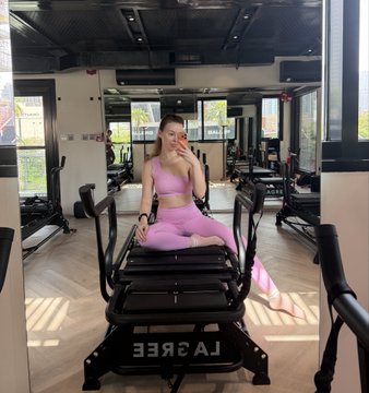 A woman in a pink workout outfit poses for a mirror selfie on a piece of gym equipment, with another person in striped pants reflected below in the mirrored surface.