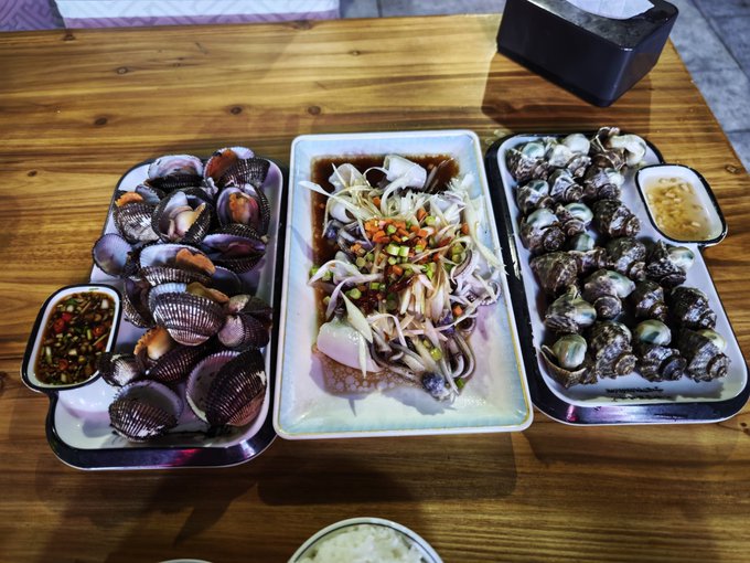 Three platters of seafood, including clams, marinated squid, and sea snails with dipping sauces, are arranged on a wooden table.