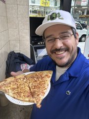 A smiling man wearing a white cap, glasses, and a blue shirt holds a paper plate with a large slice of pizza, standing outdoors with buildings visible in the background.