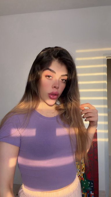 A young woman with long brown hair poses indoors in a lavender ribbed crop top, with sunlight streaming through blinds behind her.