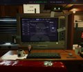 A computer monitor on a wooden desk displays code or a dark-themed interface, with a red surface visible in the foreground.
