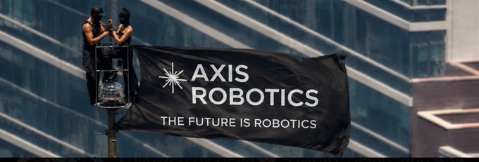 Two masked figures stand on a cherry picker displaying a black banner that reads "AXIS ROBOTICS - THE FUTURE IS ROBOTICS" against a modern glass building.