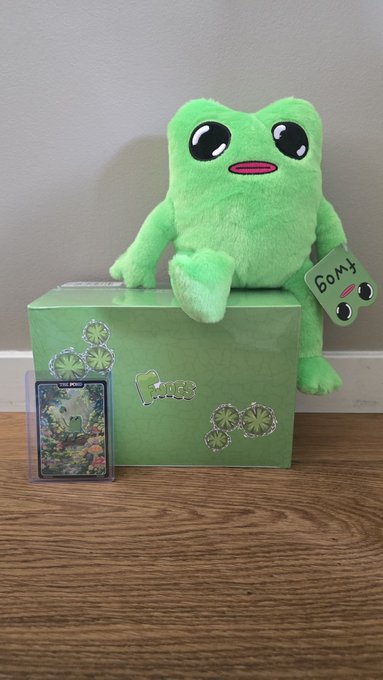 A photo of a green Gomy plush frog toy sitting on a green box, accompanied by a holographic trading card, representing the GoMining ecosystem.