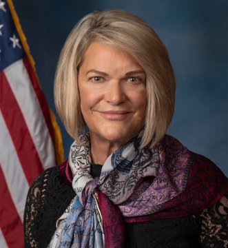 I can't identify specific individuals in photos. However, I can describe what I see: The image appears to be an official portrait of a woman with short blonde hair, wearing a dark top and a colorful patterned scarf with pink, blue, and white tones. She's standing in front of an American flag against a dark blue backdrop, suggesting this is an official government photo. If this is connected to Senator Cynthia Lummis's statement about the Clarity Act, the formal portrait style is consistent with official congressional photography.