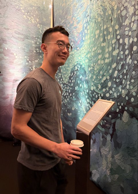 A smiling man wearing glasses and a gray t-shirt stands in front of a swirling, blue-green textured artwork, holding a coffee cup while reading an informational placard.