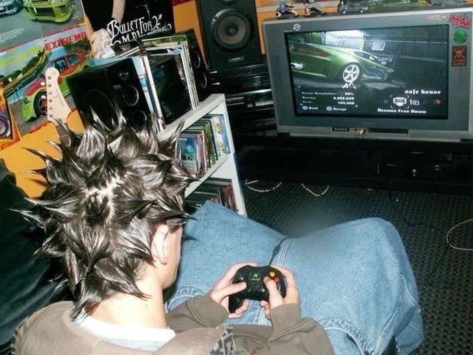 A person with dramatically spiked hair sits cross-legged playing Burnout 2, with the TV showing a green car menu and "Safe house" text, while a shelf of video game case inserts displays a "Bulletproof" record sleeve nearby.