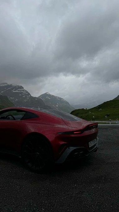 A red sports car is parked on a wet road with a scenic view of green mountains and a cloudy, overcast sky in the background.