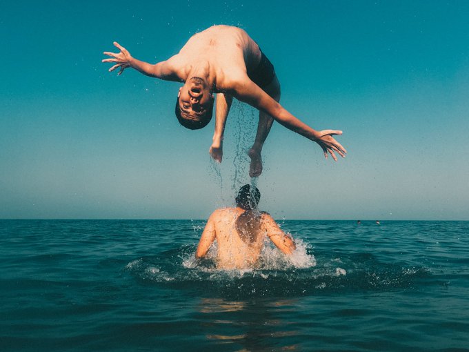 Two men in the ocean, one splashing water in the face of the other who is mid-backflip above the water.