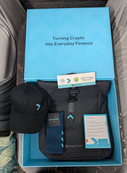 A Bitget Wallet branded merchandise box containing a black cap, backpack, and informational cards sits open with its lid reading "Turning Crypto into Everyday Finance," perfectly capturing the post's celebration of receiving this premium swag celebrating the platform's 100 million users milestone.