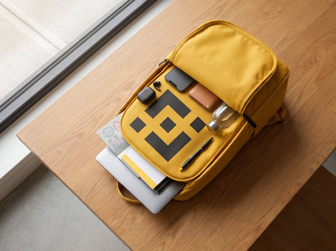 An open yellow backpack with a black crown logo on a wooden desk near a window, containing a laptop, phone, pen, AirPods case, and other items.