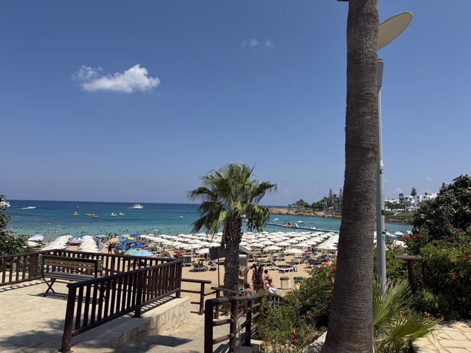 A sunny beach scene with blue water, white umbrellas, palm trees, and people enjoying the shore under a clear blue sky.