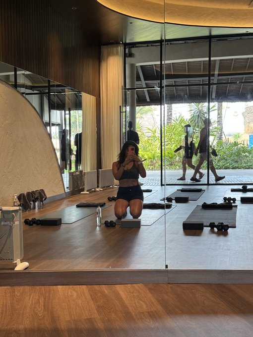 A mirror selfie of a woman kneeling on a yoga mat in a modern gym with floor-to-ceiling windows; she is holding a phone to take the photo, with exercise equipment and other people visible in the reflection.
