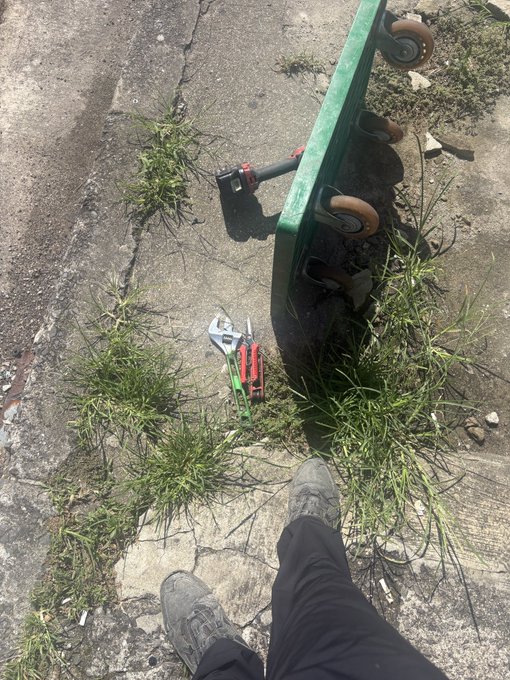 An overhead photo showing a person's booted feet standing on cracked concrete next to a tipped-over green skateboard and a discarded toy gun with red and green details, surrounded by weeds.