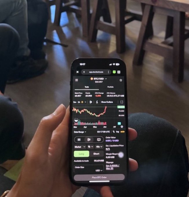 The image shows a person holding an iPhone screen displaying a cryptocurrency trading app, which seems to be showing Bitcoin information. The phone's wallpaper also displays "@AptosLabs". This suggests that the person might be interested in or involved in the world of cryptocurrencies and potentially investing in them using the trading app on their phone.