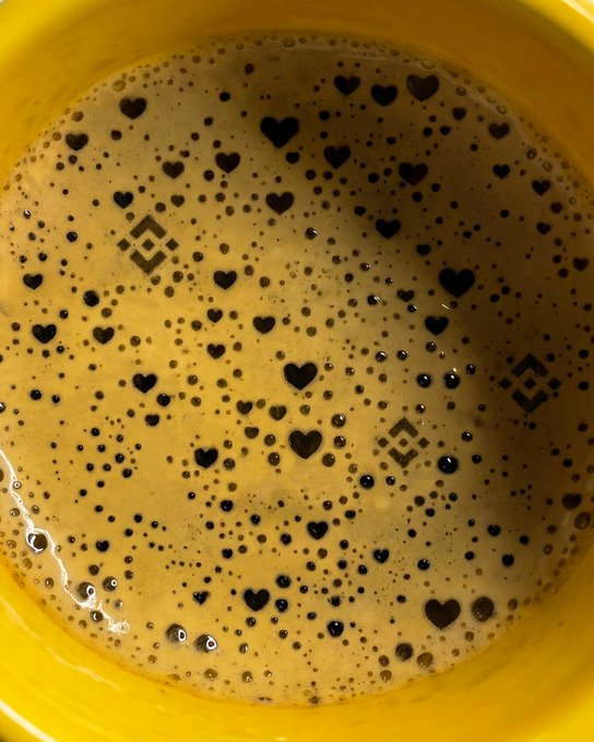 A frothy yellow beverage surface dotted with dark heart and Binance logo-shaped foam patterns sits in a yellow mug, playfully evoking the love and brand identity celebrated alongside Binance's restored Terra withdrawal capabilities.