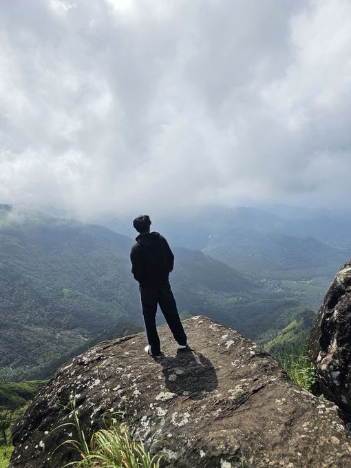 A person stands on a rocky cliff edge, gazing out over misty green mountains under a cloudy sky.