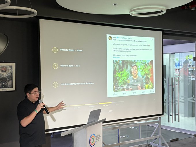 A man presenting in front of a projection screen showing a slide titled 'Direct to Wallet' with bullet points and an embedded tweet.
