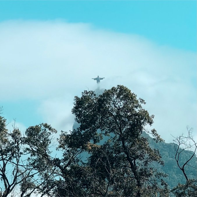 A scenic photograph of the Christ the Redeemer statue in Rio de Janeiro viewed through tree branches and misty clouds against a blue sky.