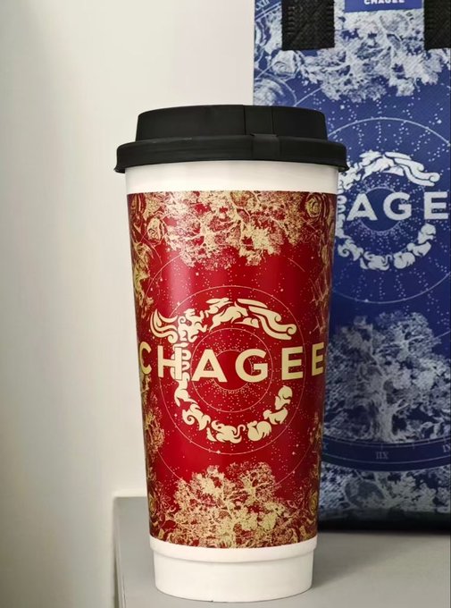 A red Chagee branded paper coffee cup with gold decorative designs sitting next to a blue Chagee shopping bag on a white surface.