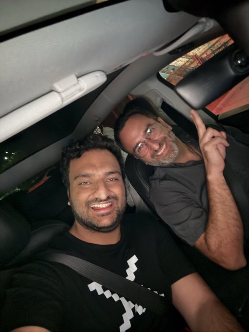 Two men taking a selfie inside a car at night; the younger man with dark curly hair and a beard smiles at the camera, while the older man with glasses behind him grins and gestures playfully.