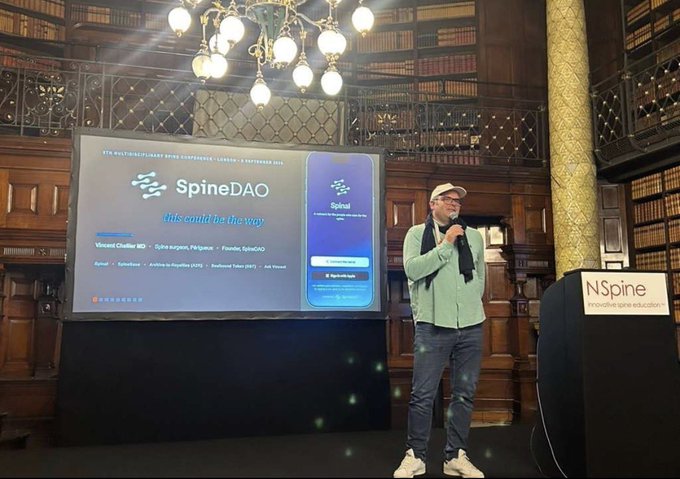 A man in a green shirt and cap speaks at a 'SpineDAO' event in an ornate library setting, with a screen displaying 'This could be the way' and 'Spinal', and an 'N-Spine' podium in the foreground.