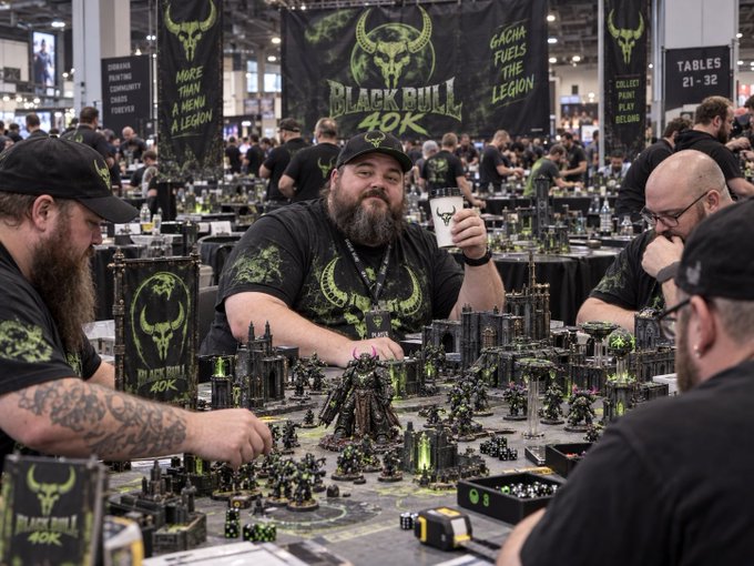 Men wearing 'Black Bull 40K' t-shirts play a Warhammer 40K tabletop game, with banners reading 'More Than a Hobby, A Legion,' 'Gacha Fuels the Legion,' and 'Tables 21-32.'