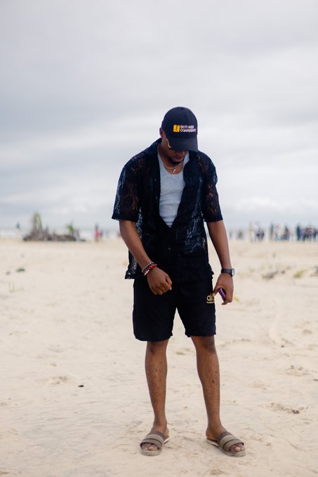 A man stands barefoot on a sandy beach wearing slide sandals, black shorts, an open black lace shirt over a grey tank top, and a black cap with 'NOMADZ' branding on it.