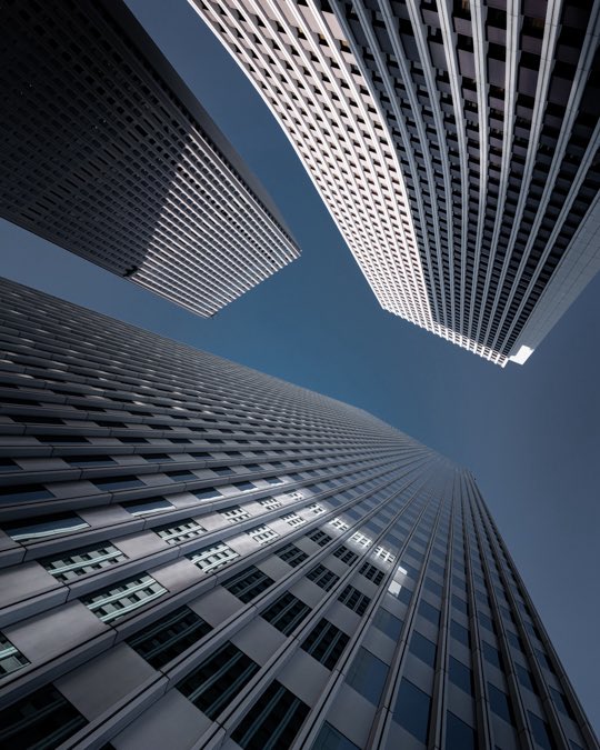 A dramatic upward-angled photograph of modern skyscrapers converging against a clear blue sky, emphasizing geometric lines and architectural perspective.