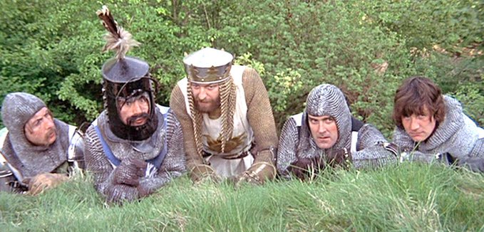 Five medieval knights in chainmail and armor peer over a grassy hill, including one wearing a crown, reminiscent of a scene from Monty Python and the Holy Grail.